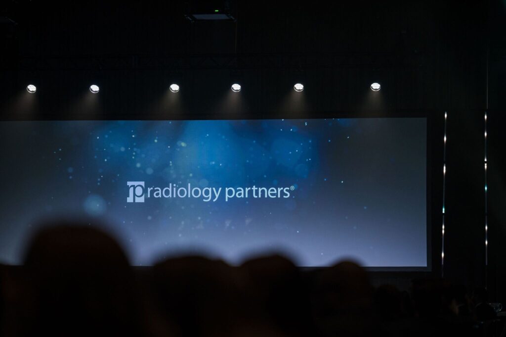 an audience faces an empty stage with a blue screen that says Radiology Partners