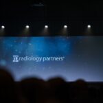 an audience faces an empty stage with a blue screen that says Radiology Partners