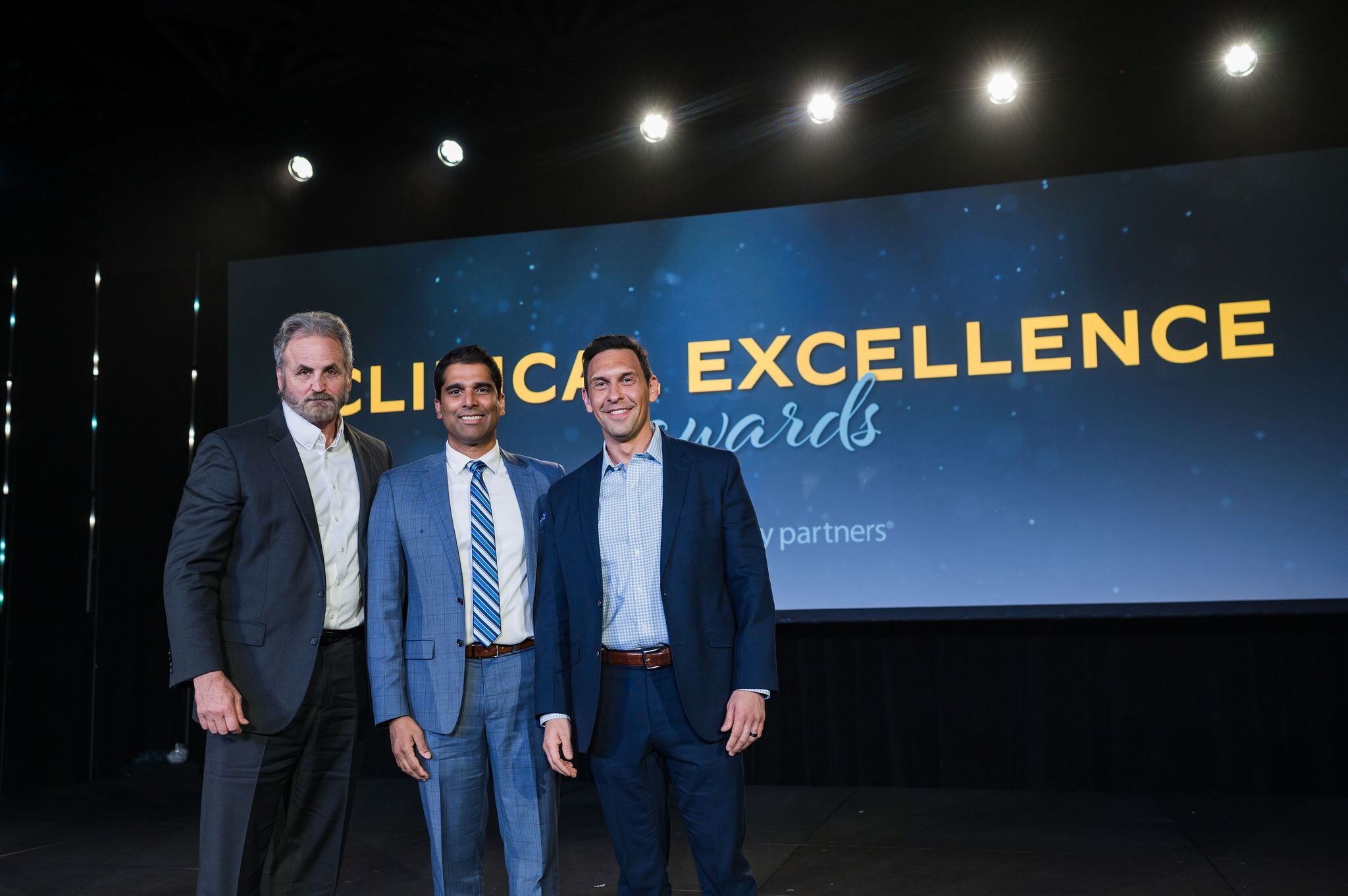 Three men stand on a stage in front of a screen that says Clinical Excellence Awards