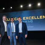 Three men stand on a stage in front of a screen that says Clinical Excellence Awards