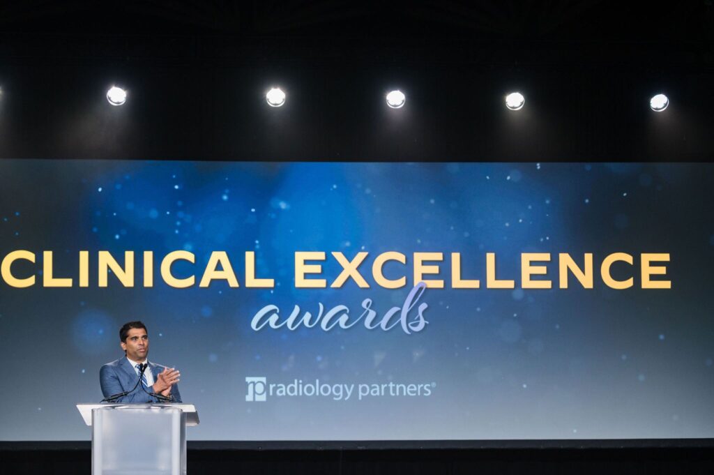 a man stands on stage at a podium clapping in front of a screen that says clinical excellence awards