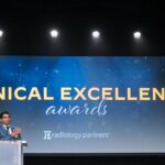 a man stands on stage at a podium clapping in front of a screen that says clinical excellence awards