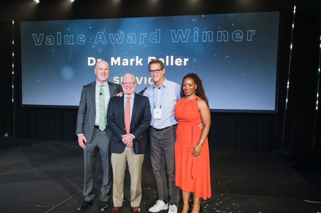 three men and one woman stand on stage smiling