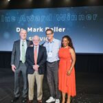 three men and one woman stand on stage smiling