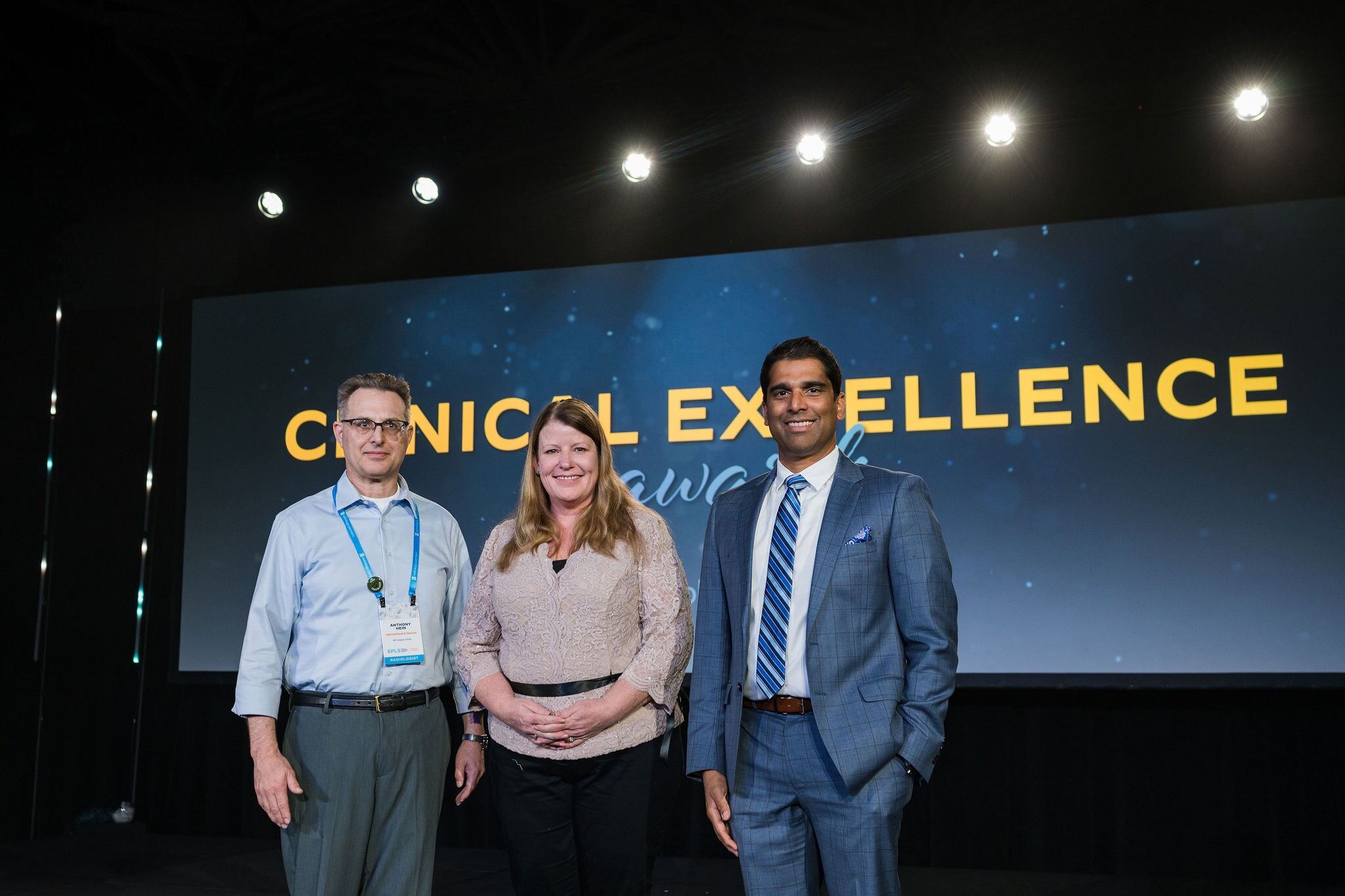 two men and one woman stand on stage in front of a screen that says clinical excellence awards