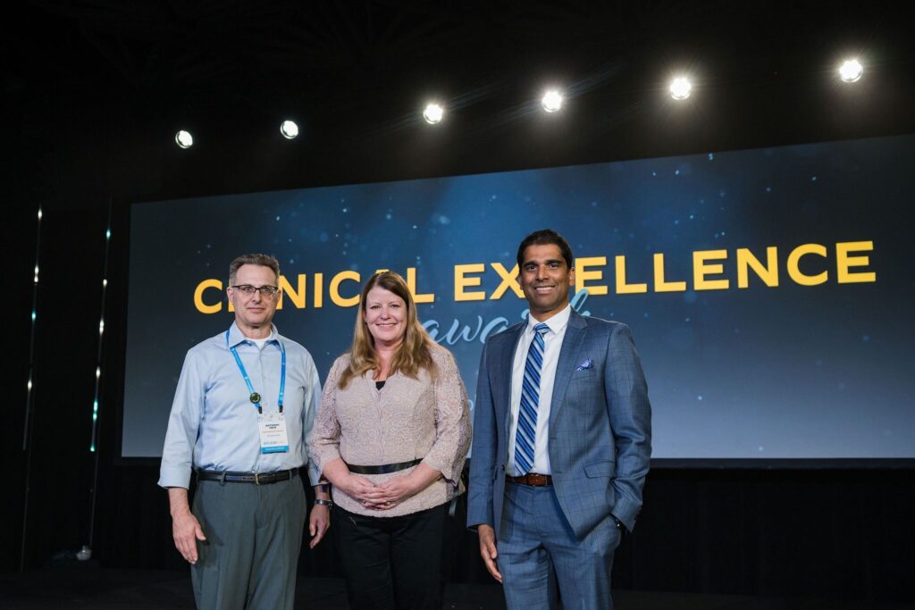 two men and one woman stand on stage in front of a screen that says clinical excellence awards