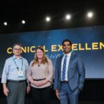 two men and one woman stand on stage in front of a screen that says clinical excellence awards