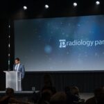 a man stands on stage speaking at a podium in front of a blue screen that says radiology partners