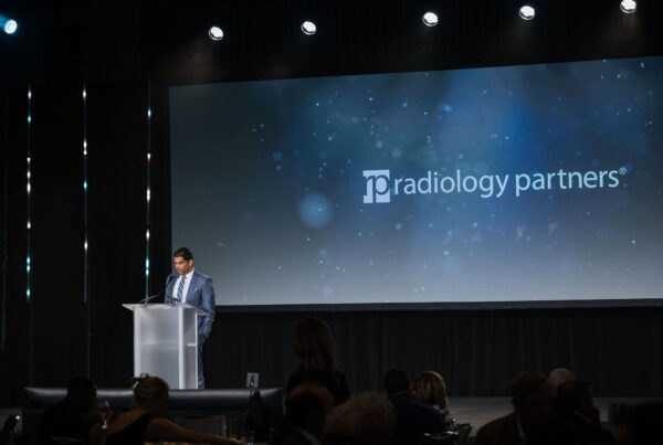 a man stands on stage speaking at a podium in front of a blue screen that says radiology partners