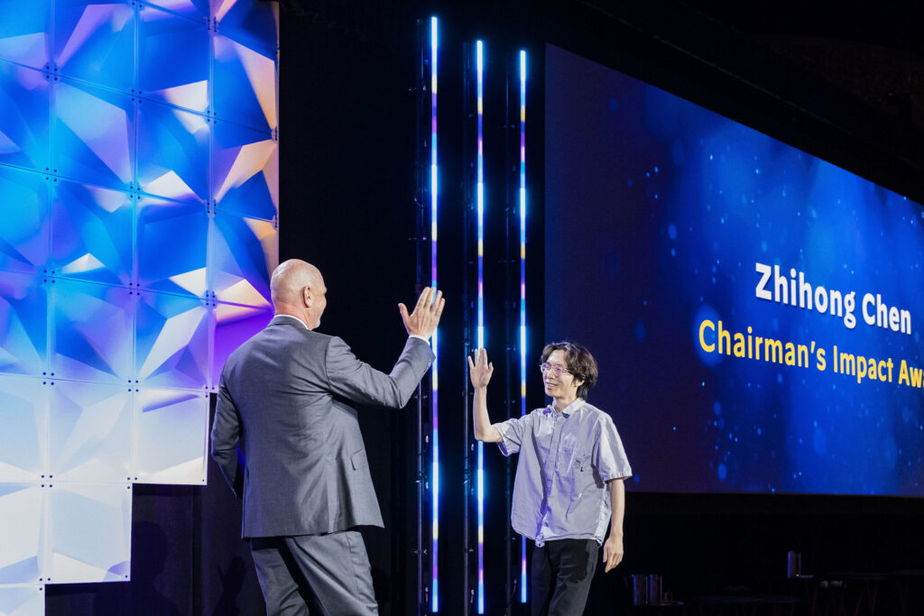 A man in a suit and a man in a short-sleeved shirt high-five on stage at an awards event. A large screen behind them displays Zhihong Chen and Chairmans Impact Award.