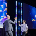 A man in a suit and a man in a short-sleeved shirt high-five on stage at an awards event. A large screen behind them displays Zhihong Chen and Chairmans Impact Award.