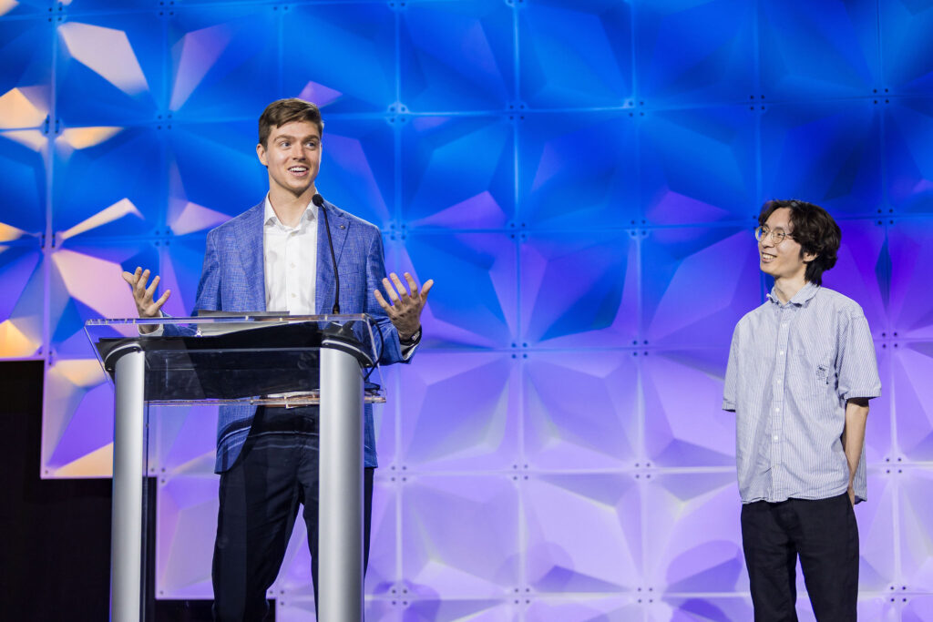 Two people stand on stage with a geometric blue and purple backdrop. One person in a blazer speaks at a clear podium, gesturing with hands, while the other in a short-sleeved shirt stands and looks at him, smiling.