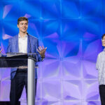 Two people stand on stage with a geometric blue and purple backdrop. One person in a blazer speaks at a clear podium, gesturing with hands, while the other in a short-sleeved shirt stands and looks at him, smiling.