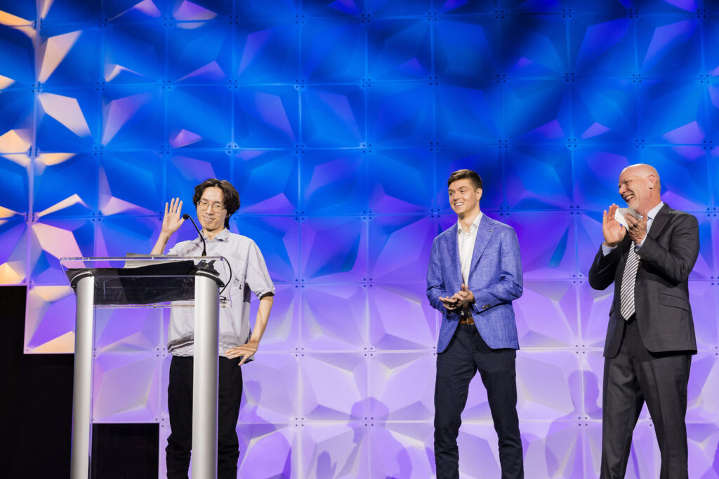 A person stands at a podium waving while two others stand nearby, smiling and applauding, against a geometric blue and purple-lit background on a stage.