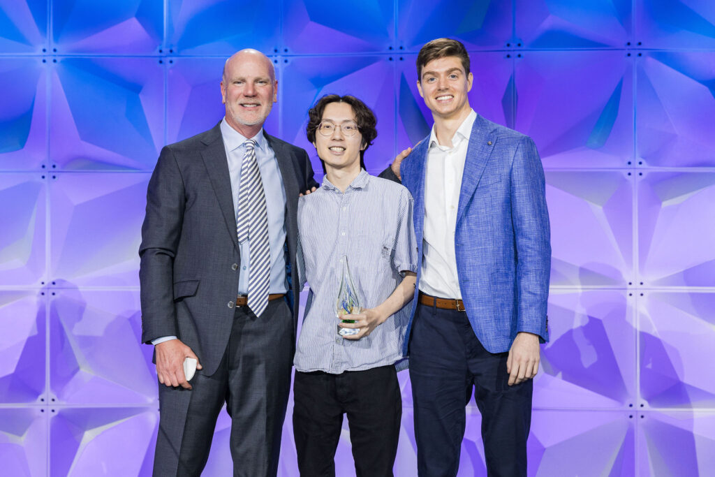 Three men stand together smiling in front of a geometric purple backdrop. The man in the middle holds a glass award. All are dressed in business attire, with jackets and collared shirts.