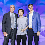 Three men stand together smiling in front of a purple geometric backdrop. The man in the center holds a glass award. All are dressed in business attire, with the two men on each side wearing suits and the center man in a shirt and pants.