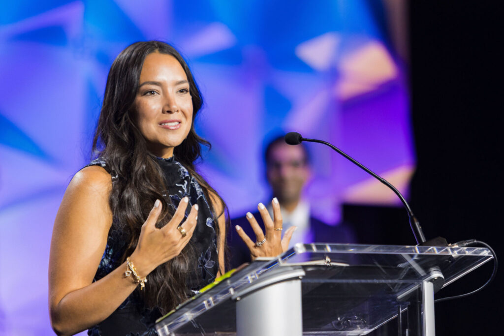 A woman with long dark hair speaks at a clear podium with a microphone, gesturing with her hands. The background is blue and purple with abstract shapes, and a blurred person stands behind her.