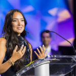 A woman with long dark hair speaks at a clear podium with a microphone, gesturing with her hands. The background is blue and purple with abstract shapes, and a blurred person stands behind her.