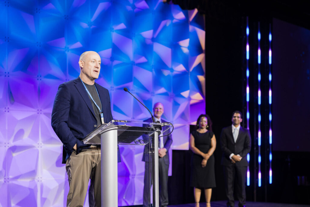 A man speaks at a clear podium on a stage with a geometric blue and purple background, while three people stand behind him, two men and one woman, all dressed in formal attire.