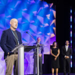 A man speaks at a clear podium on a stage with a geometric blue and purple background, while three people stand behind him, two men and one woman, all dressed in formal attire.