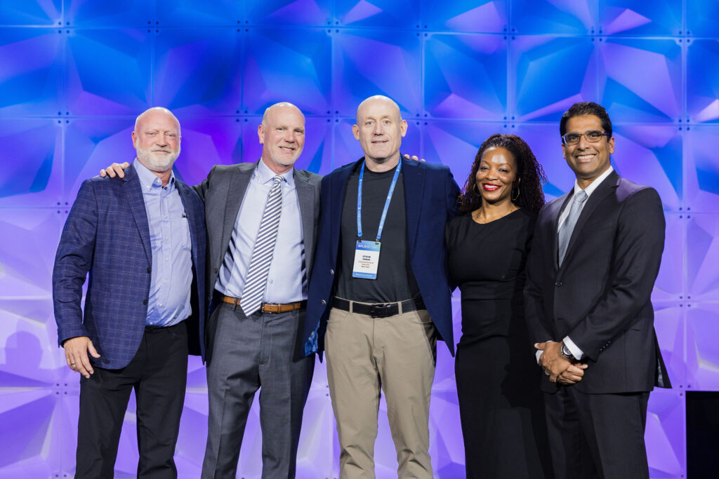 Five people in business attire stand together smiling on stage in front of a blue and purple geometric backdrop at a professional event or conference.