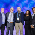 Five people in business attire stand together smiling on stage in front of a blue and purple geometric backdrop at a professional event or conference.