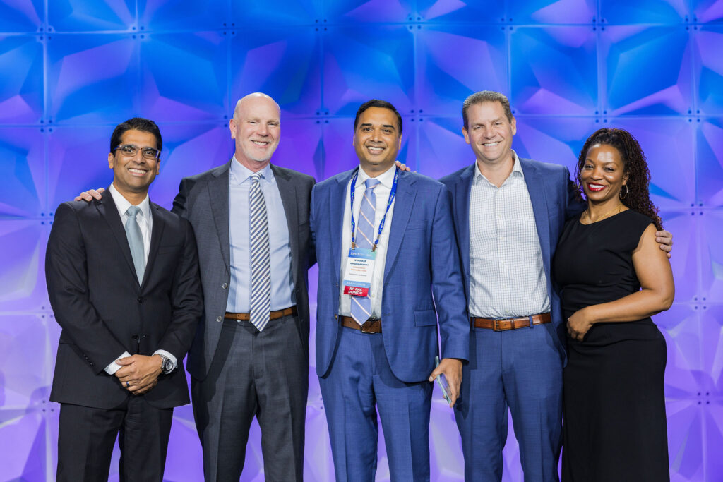Five professionally dressed people stand smiling in front of a blue geometric backdrop at an event. Three men wear suits and ties, one man is in a jacket, and one woman is in a black dress.