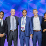 Five professionally dressed people stand smiling in front of a blue geometric backdrop at an event. Three men wear suits and ties, one man is in a jacket, and one woman is in a black dress.