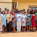 group of about 20 people, men and women, are posing and smiling together outside a school