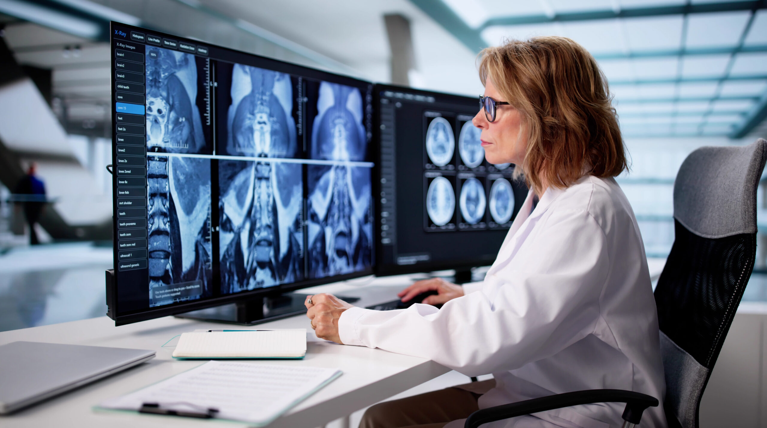 A female doctor in a white lab coat sits at a desk analyzing spinal and brain MRI scans for a teleradiology practice, with advanced imaging services displayed on large monitors in a modern medical office.